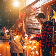 Eine Frau und ein Mann halten gemeinsam eine Lichterkette mit Kugeln fest. Die Menschen stehen vor einem Holzhaus und sind weihnachtlich gekleidet.