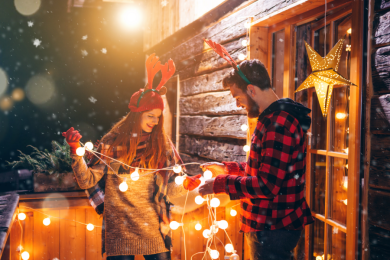 Eine Frau und ein Mann halten gemeinsam eine Lichterkette mit Kugeln fest. Die Menschen stehen vor einem Holzhaus und sind weihnachtlich gekleidet.