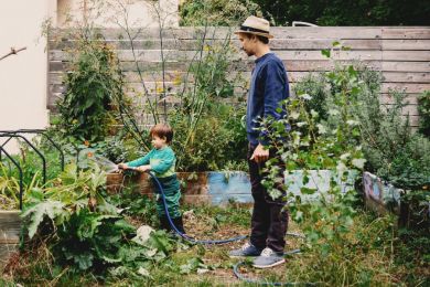 Ein Junge steht mit seinem Vater im Garten und sprengt ein Hochbeet mit Regenwasser.