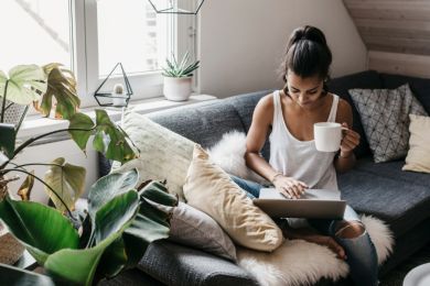 Junge Frau sitzt auf der Couch mit einer Tasse Kaffee und einem Laptop.