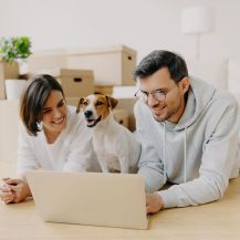 Ein junges Paar liegt mit seinem Hund auf dem Wohnzimmerfußboden vor gepackten Umzugskartons.