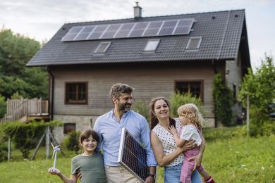 Eine junge Familie vor Ihrem Haus mit Solarpanelen auf dem Dach. 