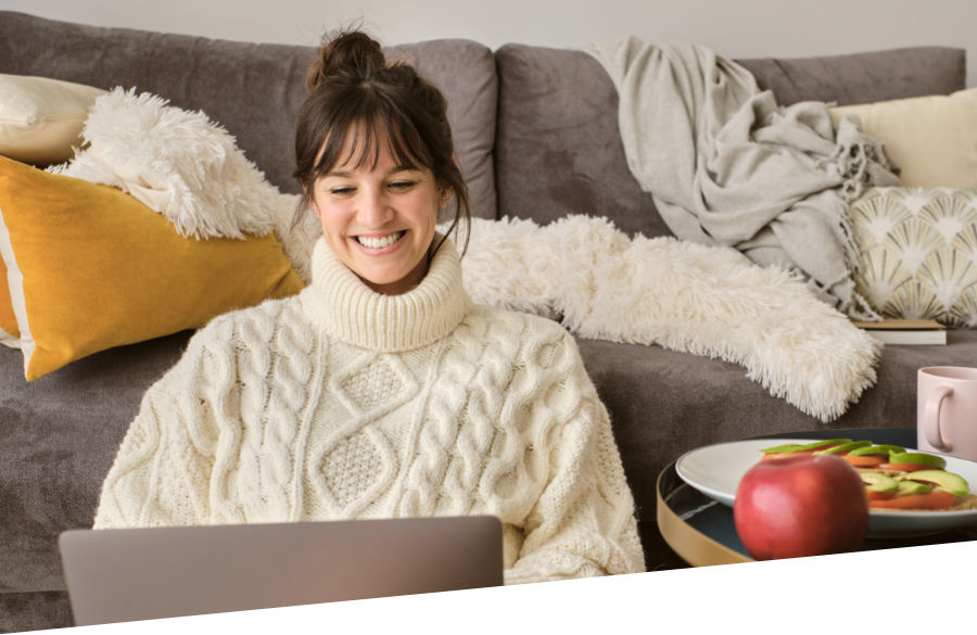 Eine Frau mit einem warmen Rollkragenpullover sitzt auf dem Boden und lehnt sich an ein Sofa. Sie nutzt ihr Notebook und lacht dabei fröhlich.